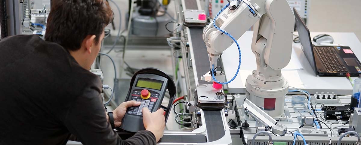 Image-Man is holding teach panel to control a robotic arm which is integrated on smart factory production line.