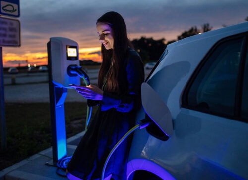 Young lady on phone charging her electric car at a charging station