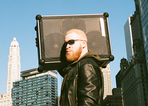 Image-Cool young man wearing sunglasses carrying a Soundboks speaker on his shoulder and skyscrapers in the background.